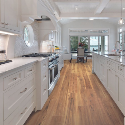  A bright, coastal-style kitchen with medium-brown wood-look flooring, white cabinetry, and a scenic outdoor view through large windows.